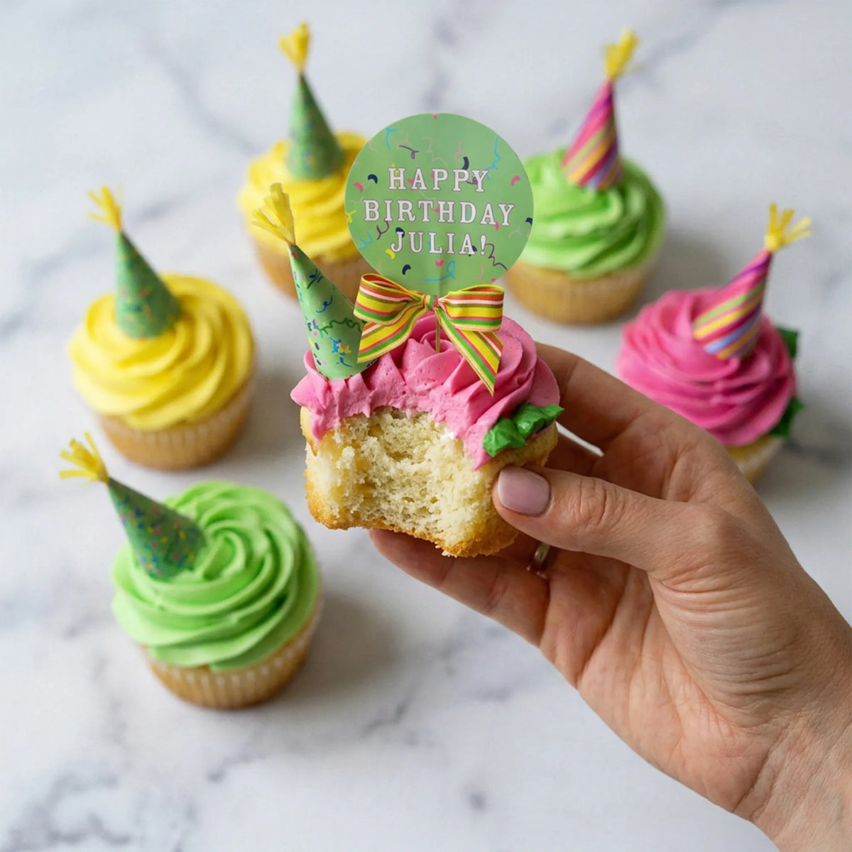 Hand holding a bitten vanilla party hat cupcake with pink buttercream and a Happy Birthday topper, kosher, made in Chestnut Hill MA