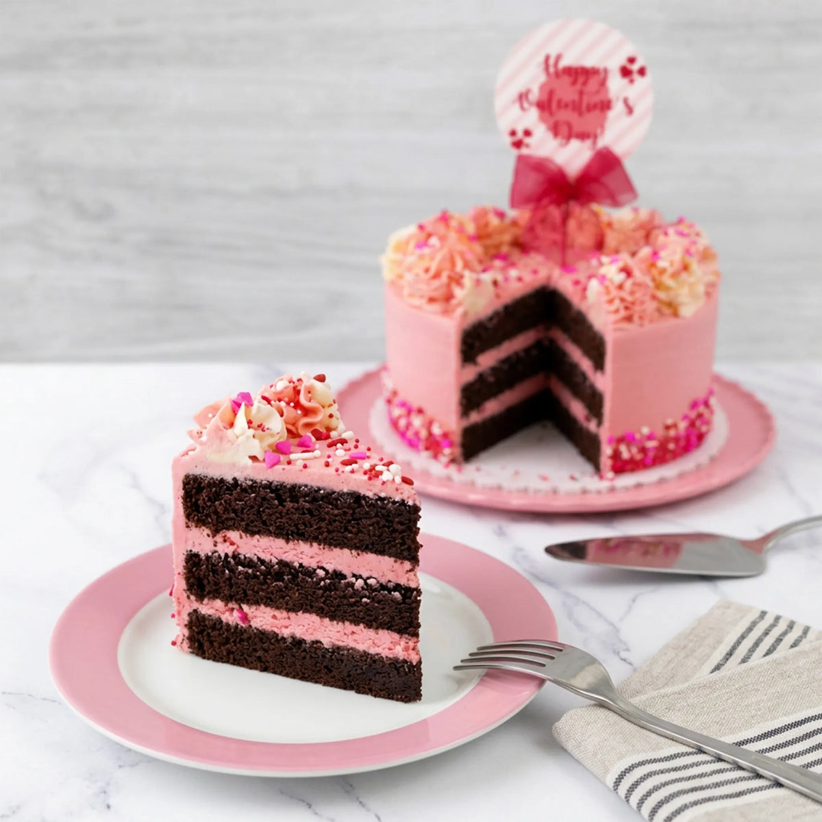 Slice of Valentine's Day sprinkles cake showing chocolate layers with pink buttercream, with the cut cake behind it, kosher, made in Chestnut Hill MA