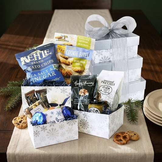 Large Holiday Gift Tower option 2: stacked silver snowflake boxes with a sheer silver bow and open boxes of Kettle chips, cookie bites, seasoned pretzels and foil truffles, styled on a walnut table with a linen runner and pine sprigs