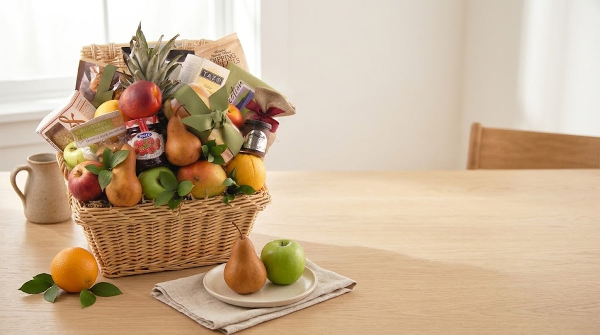 Fresh fruit and gourmet gift basket with pears, apples, oranges, a pineapple, TAZO tea, Hero raspberry preserves and dipping pretzels on a pale oak dining table, set out for an everyday just because gift