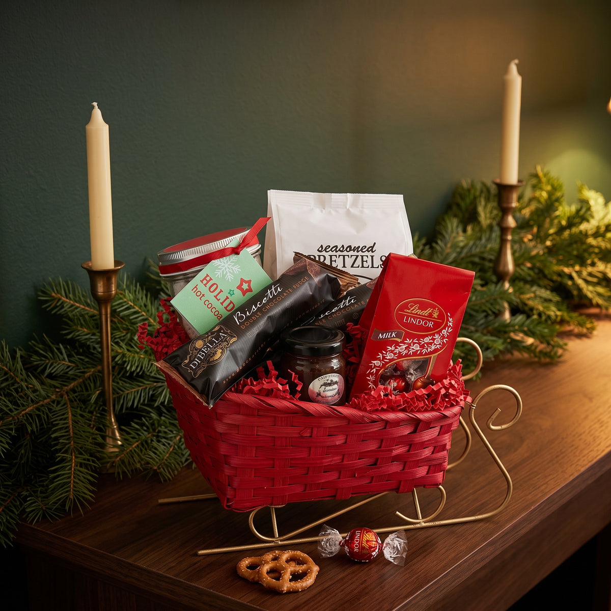 Holiday sleigh gift basket on gold runners filled with pretzels, cocoa, biscotti and Lindt chocolates, styled on a wood mantel with spruce and candles