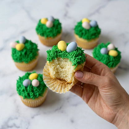 Hand holding a bitten vanilla Easter cupcake with buttercream grass and pastel candy eggs, kosher, made in Chestnut Hill MA