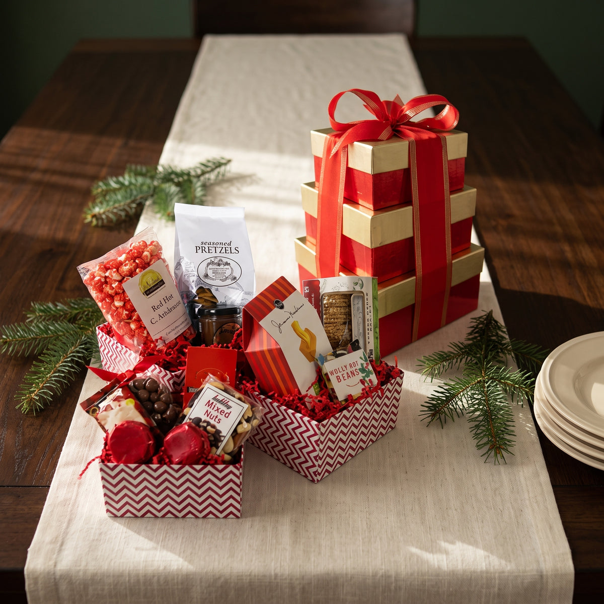 Large holiday gift tower of stacked red and gold boxes with pretzels, popcorn, nuts and truffles, styled on a linen runner along a dining table