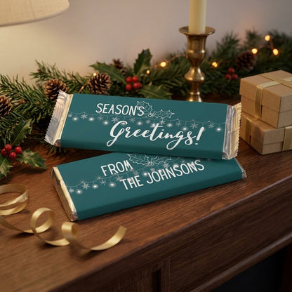 Season's Greetings design, wrapped Hershey bars in pine green with snowflake lights, styled on a wood mantel with evergreen garland, holly and wrapped gifts