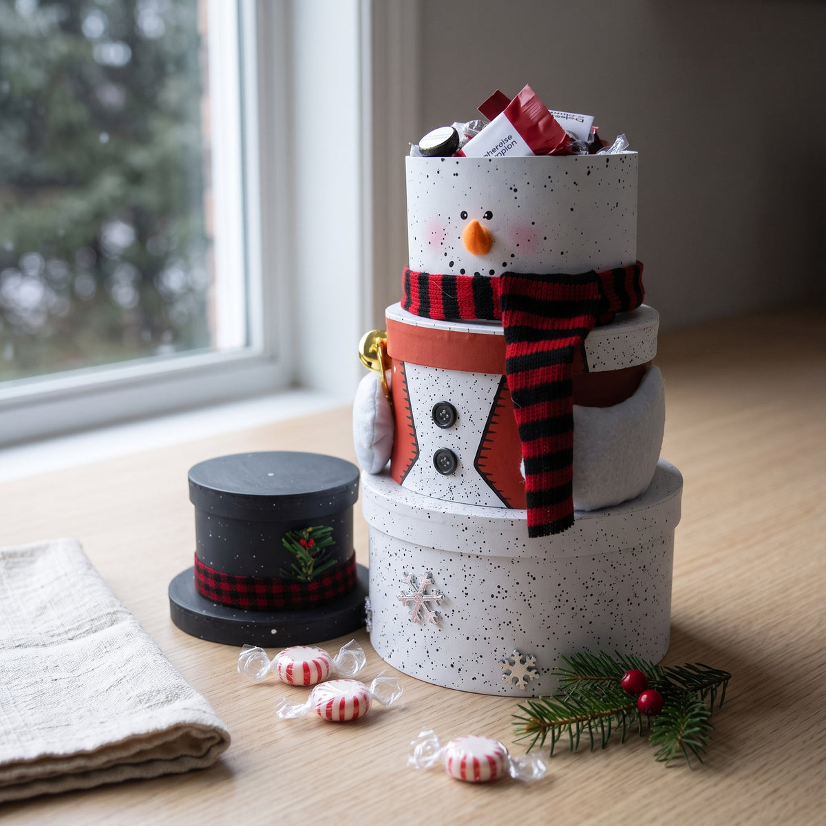 Snowman gift tower of four stacked round boxes with a buffalo check scarf and top hat lid, styled on a kitchen counter with peppermints and spruce