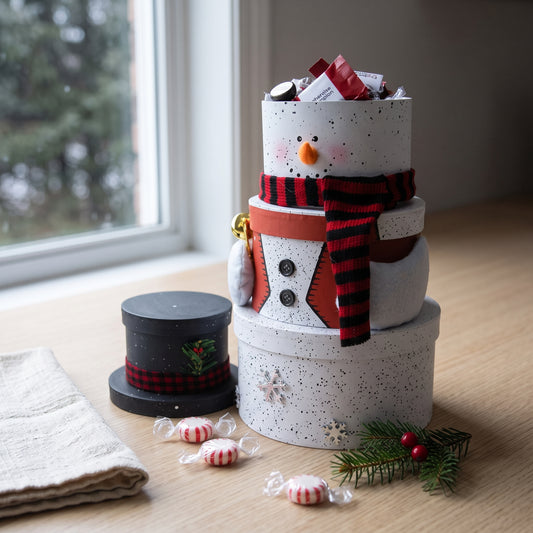 Snowman gift tower of four stacked round boxes with a buffalo check scarf and top hat lid, styled on a kitchen counter with peppermints and spruce